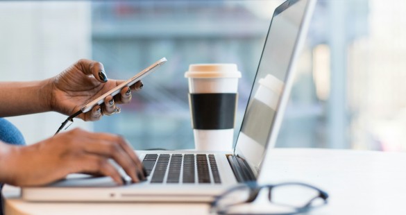 Person using a smartphone and laptop at a desk with a coffee cup, representing multitasking and productivity in a modern workspace.